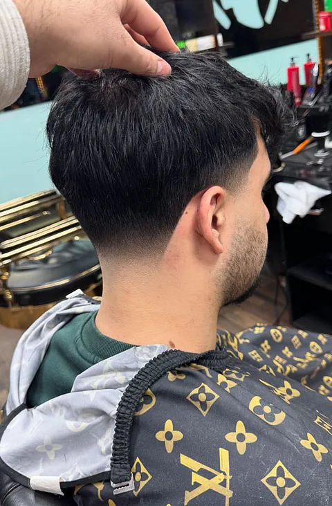 Mann mit kurzem, gestuften Haarschnitt, der von hinten betrachtet wird. Er trägt einen Umhang mit Louis Vuitton-Muster. Eine Hand greift nach seinem Haar.